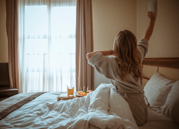 Giovane donna seduta sul letto allungando la mano con la colazione sul tavolo