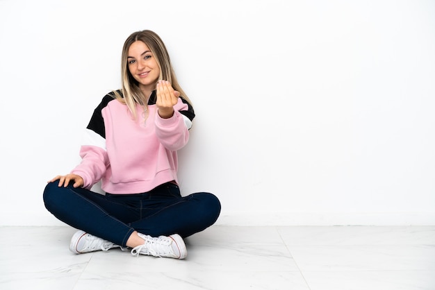 Giovane donna seduta sul pavimento al chiuso che invita a venire con la mano. Felice che tu sia venuto