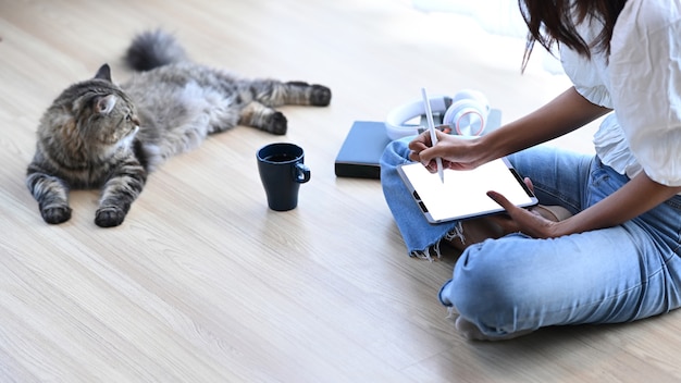 Giovane donna seduta sul pavimento con il suo gatto e lavorando su tavoletta digitale nel soggiorno di casa.