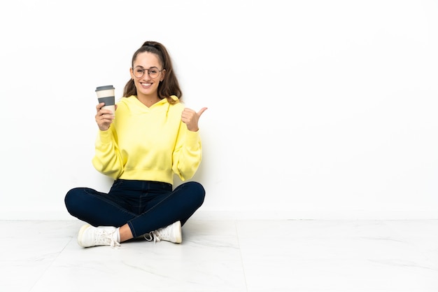 Giovane donna seduta sul pavimento con in mano un caffè da asporto che punta di lato per presentare un prodotto