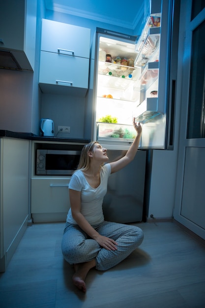 Giovane donna seduta sul pavimento e che prende il cibo dal frigorifero aperto a tarda notte