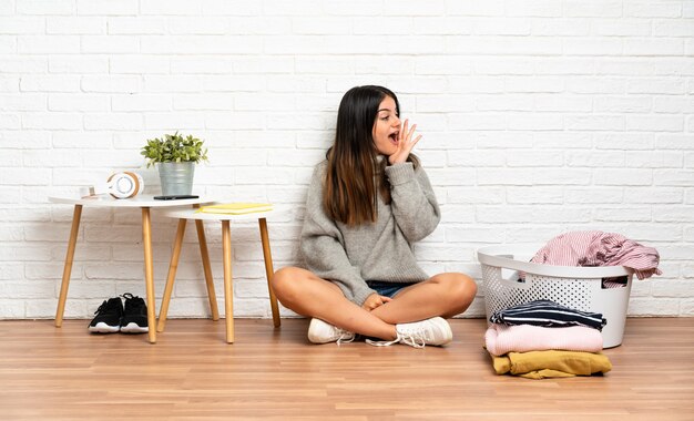 Giovane donna seduta sul pavimento in casa con cesto di vestiti che grida con la bocca spalancata al lato