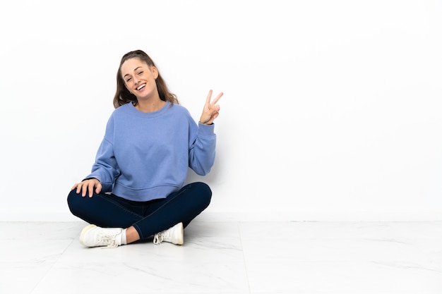 Giovane donna seduta sul pavimento sorridendo e mostrando il segno di vittoria