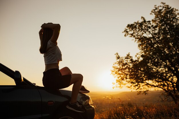 Giovane donna seduta sulla macchina al tramonto. Si gode il bellissimo paesaggio e il tramonto.