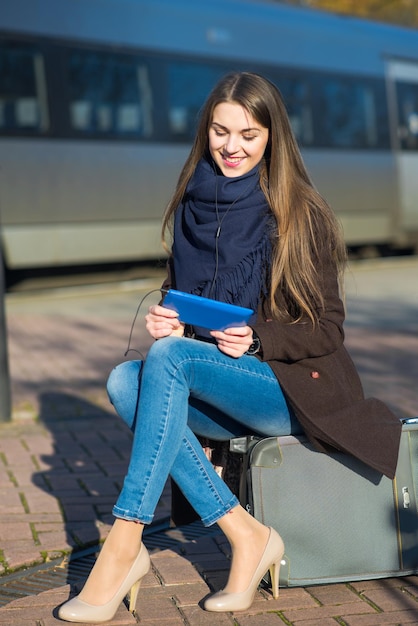 Giovane donna seduta sulla valigia utilizzando un tablet alla stazione ferroviaria