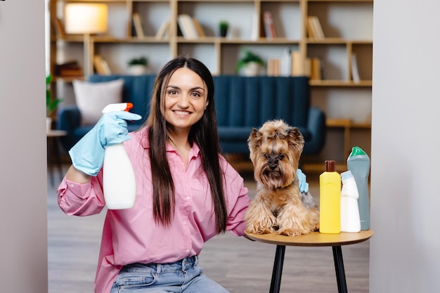 Giovane donna seduta vicino al tavolo con prodotti per la pulizia a casa e anche il suo cagnolino è seduto sul tavolo