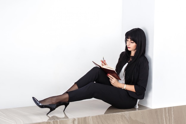 Giovane donna seria attraente che si siede su un pavimento in un ufficio, leggente in un taccuino.