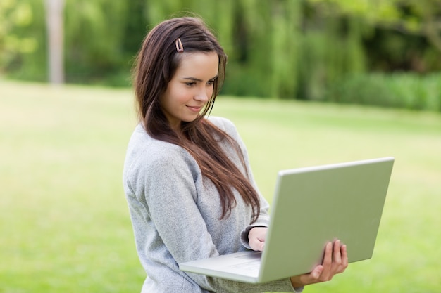 Giovane donna seria che sta dritta in un parco mentre tenendo il suo computer portatile