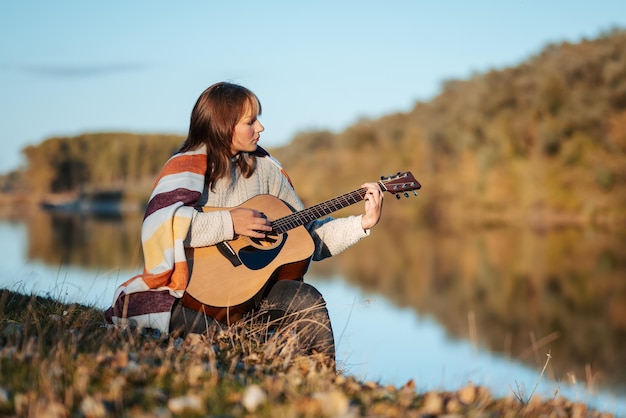 Giovane donna seria che suona la chitarra sulla riva del fiume concetto di godersi la sera soleggiata in natura ora d'oro tempo fresco e musica