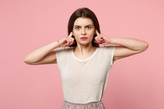 Giovane donna seria in abiti leggeri casual in posa isolata su sfondo rosa pastello parete ritratto in studio. Persone sincere emozioni concetto di stile di vita. Mock up spazio di copia. Coprire le orecchie con le dita.