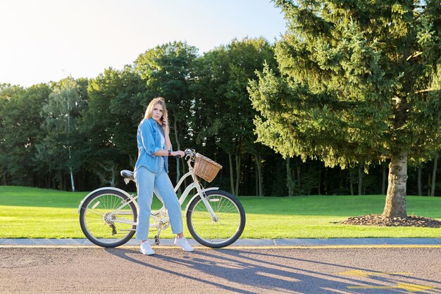 Giovane donna seria sulla strada con la bici che parla sul telefono, fondo dell'erba del parco verde, tramonto di giorno soleggiato, spazio della copia