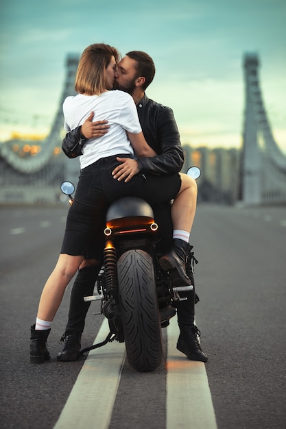 Giovane donna sexy che abbraccia uomo carino in giacca di pelle nera elegante, seduto su moto sportiva sul ponte della città al tramonto e baciare.