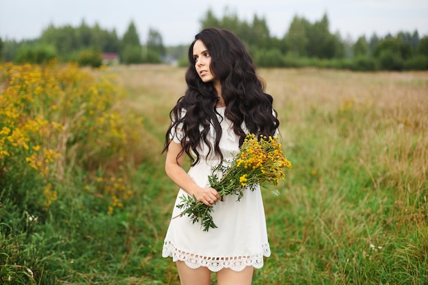 Giovane donna sexy del brunette in breve vestito bianco che cammina in un campo di fioritura. Ragazza di modello dolce in breve vestito rustico che tiene i fiori freschi del campo e che posa su un prato della molla. Natura di primavera. Estate