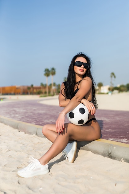 Giovane donna sexy in forma in occhiali da sole che tiene il pallone da calcio sulla spiaggia