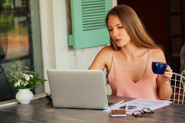 Giovane donna sexy in una camicia elegante utilizzando laptop e telefono freelance sulla spiaggia paradiso tropicale Ragazza tazza di tè caffè lavoro freelance seduto in un caffè estivo lavoro a distanza Oceano Indiano