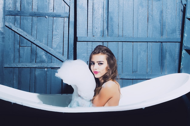 Giovane donna sexy nella vasca da bagno bianca che gioca con la schiuma di sapone