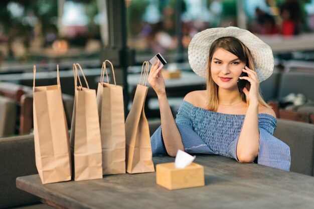 Giovane donna shopaholic allegra che si siede in caffè all'aperto mentre parlando al telefono e tenendo la carta di credito in sua mano