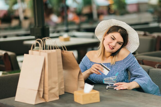 Giovane donna shopaholic allegra online con i sacchi di carta di acquisto che si siedono in caffè all'aperto e che tengono la carta di credito e mini carrello di acquisto in sue mani mentre parla sul telefono