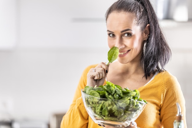 Giovane donna sicura che tiene una ciotola piena di spinaci freschi Tiene una foglia sul viso Concetto di stile di vita sano