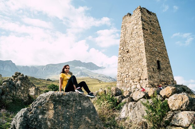 Giovane donna siede su pietra vicino a vecchie torri sullo sfondo di possenti montagne avvolte in nuvole Turista che riposa accanto a edifici storici distrutti dopo un attivo trekking in montagna