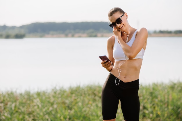 Giovane donna slim fit con auricolari e smartphone ascolta musica nel parco durante la giornata di sole