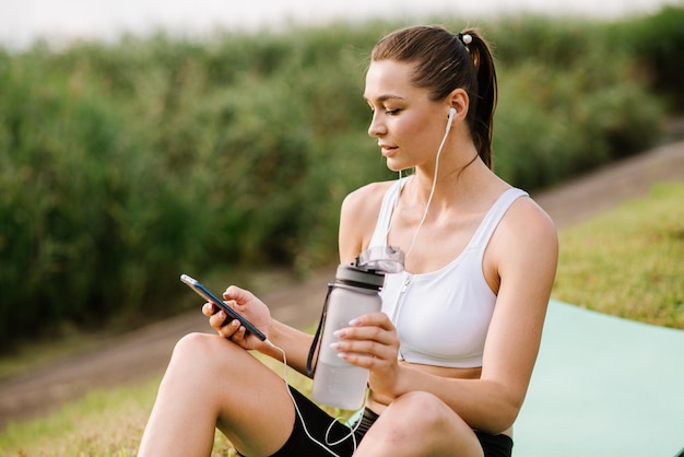 Giovane donna slim fit con auricolari e smartphone ascolta musica nel parco durante la giornata di sole