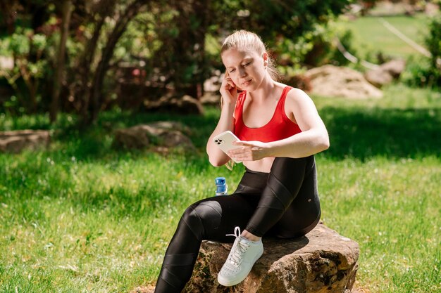 Giovane donna slim fit in abbigliamento sportivo che riposa utilizzando il cellulare dopo gli allenamenti nel parco