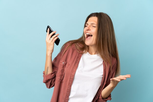 Giovane donna slovacca isolata su sfondo blu utilizzando il telefono cellulare e cantando