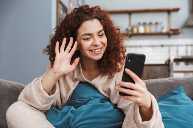 giovane donna soddisfatta che indossa abiti da casa seduta sul divano in appartamento e usa il cellulare
