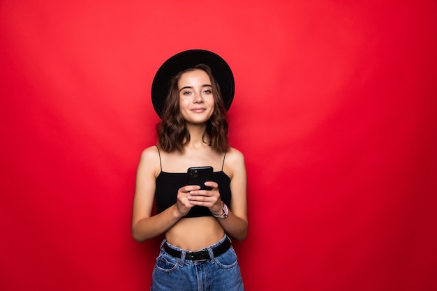 Giovane donna soddisfatta in vestiti neri che guarda sulla macchina fotografica mentre usando il telefono delle cellule con gioia sopra il rosso