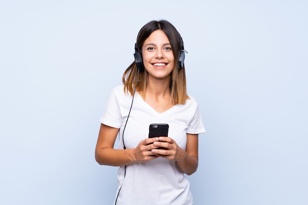 Giovane donna sopra blu isolato utilizzando il cellulare con le cuffie