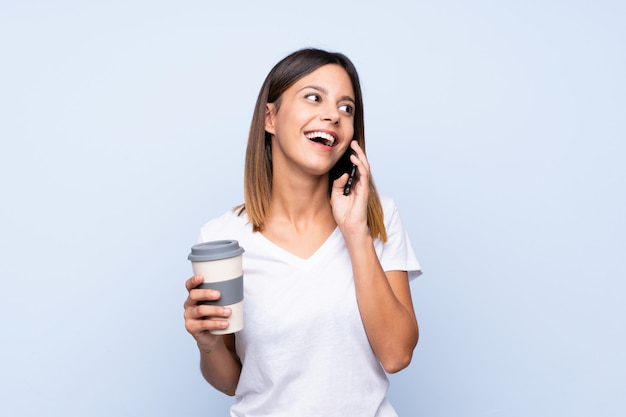 Giovane donna sopra il caffè blu isolato della tenuta da portare via e un cellulare