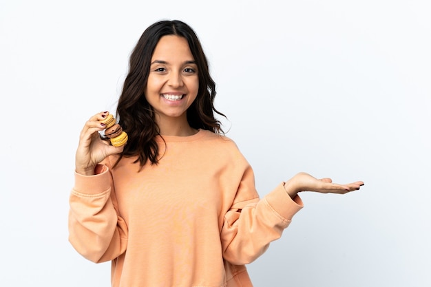 Giovane donna sopra la parete bianca isolata che tiene i macarons francesi variopinti con l'espressione scioccata