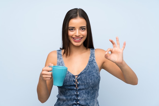 Giovane donna sopra la parete blu isolata che tiene tazza di caffè calda