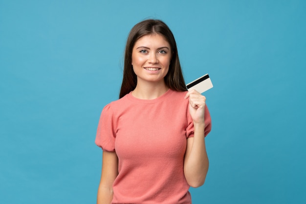 Giovane donna sopra la parete blu isolata che tiene una carta di credito