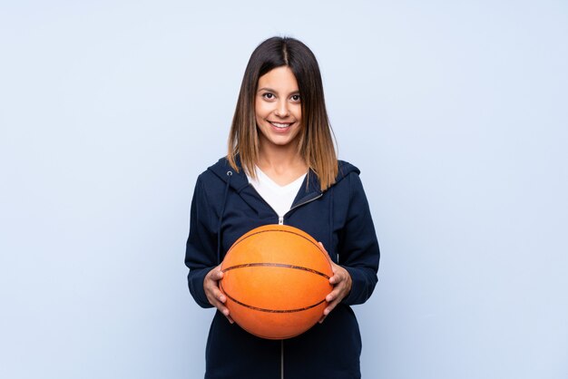 Giovane donna sopra la parete blu isolata con la palla di pallacanestro