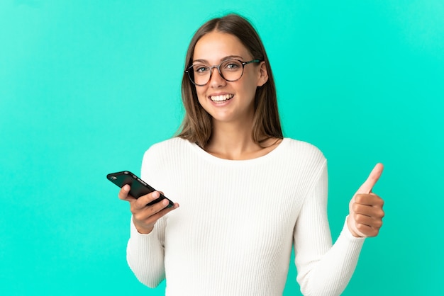 Giovane donna sopra la parete blu isolata usando il telefono cellulare mentre fa i pollici in su