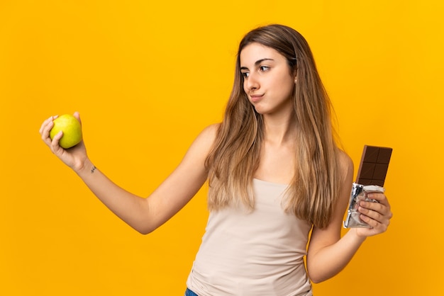 Giovane donna sopra la parete gialla isolata che ha dubbi mentre prende una compressa di cioccolato in una mano e una mela nell'altra