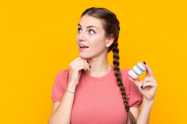 Giovane donna sopra la parete gialla isolata che tiene i macarons francesi variopinti e che pensa un'idea