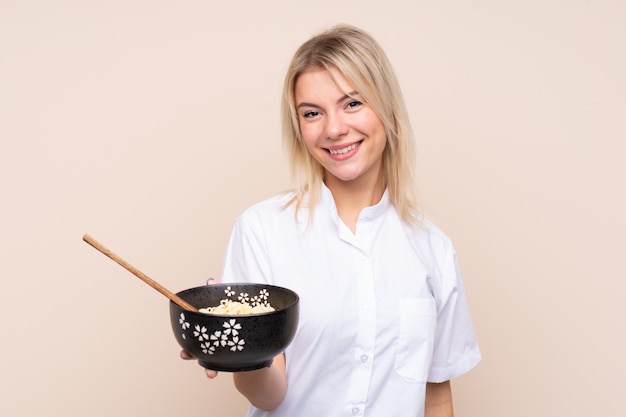 Giovane donna sopra la parete isolata con espressione felice mentre si tiene una ciotola di noodles con le bacchette