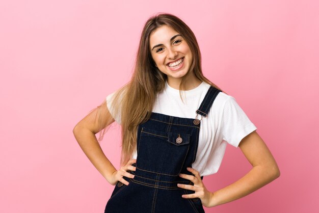 Giovane donna sopra la parete rosa isolata che posa con le braccia all'anca e sorridente