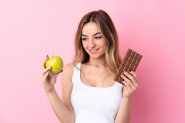Giovane donna sopra la parete rosa isolata che prende una compressa di cioccolato in una mano e una mela nell'altra