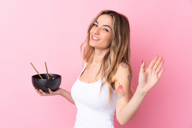 Giovane donna sopra la parete rosa isolata che saluta con la mano con l'espressione felice mentre tenendo una ciotola di tagliatelle con le bacchette