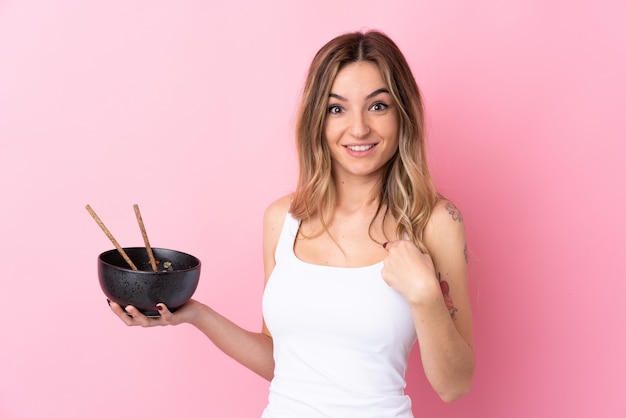 Giovane donna sopra la parete rosa isolata con espressione facciale a sorpresa mentre si tiene una ciotola di noodles con le bacchette