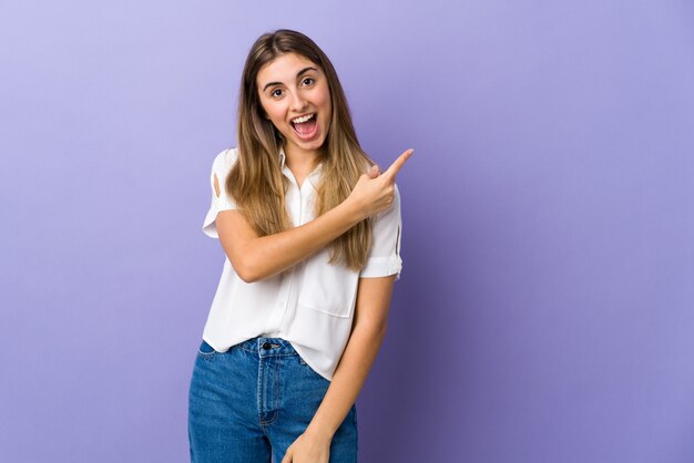 Giovane donna sopra la parete viola isolata sorpresa e indicando il lato