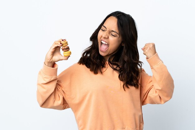 Giovane donna sopra priorità bassa bianca isolata che tiene i macarons francesi variopinti e che celebra una vittoria