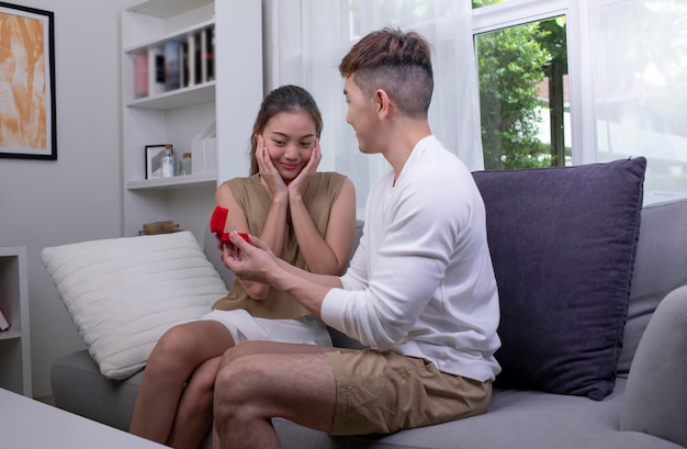 Giovane donna sorpresa con alzando le mani e sorridendo mentre il suo ragazzo la propone in soggiorno Coppia emotiva innamorata Concetto di stile di vita delle coppie asiatiche
