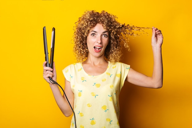 Giovane donna sorpresa con capelli ricci con piastra per capelli su sfondo giallo.