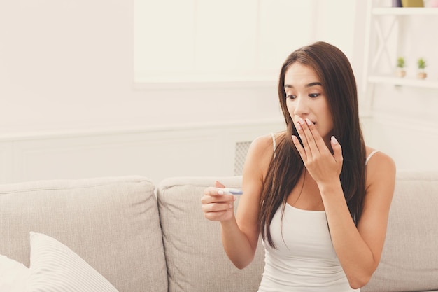 Giovane donna sorpresa con il test di gravidanza seduto sul divano a casa, concetto di maternità e aspettativa