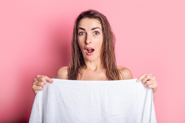 Giovane donna sorpresa con nudo di capelli bagnati che si nasconde dietro l'asciugamano su fondo rosa.